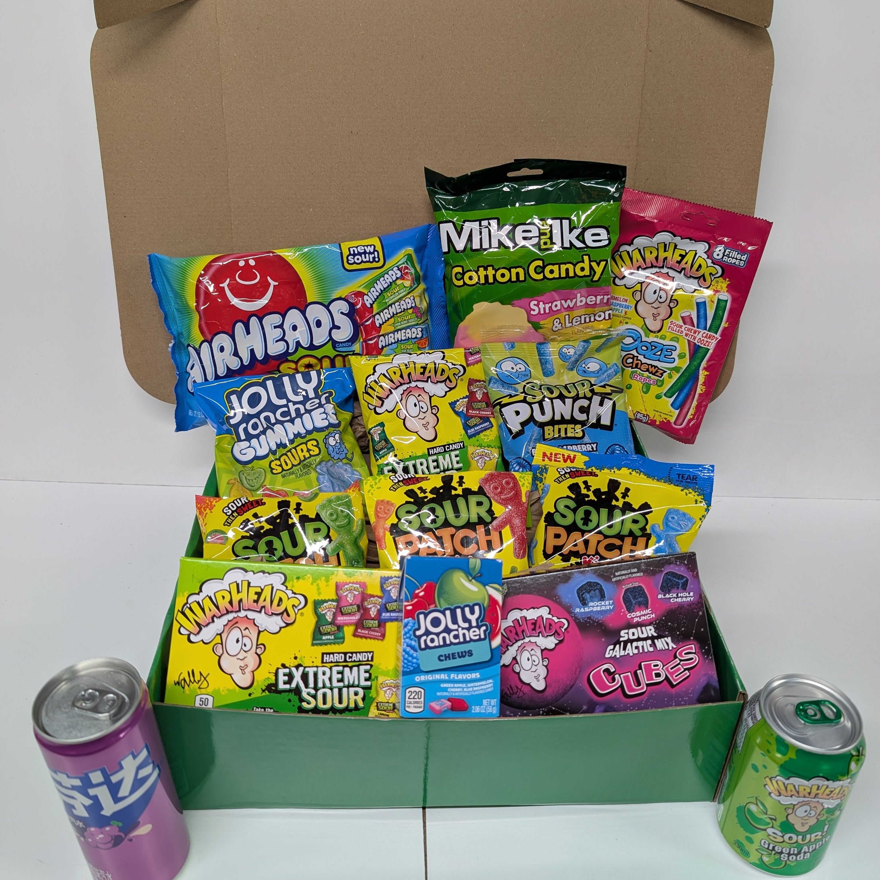 Assorted candy and soda cans in front of a cardboard box on a white background